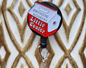 May include: A black and red enamel pin shaped like a small cooler with the text "Don't hate me because I'm a little cooler" on it. The pin is attached to a retractable badge holder.