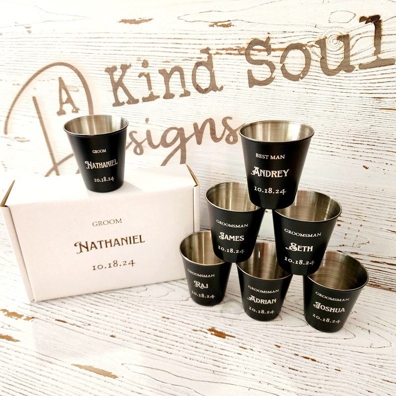 May include: Set of seven black shot glasses with gold lettering. The glasses are arranged in a pyramid shape. The glasses are engraved with the names "Nathaniel", "Andrey", "James", "Seth", "Raj", "Adrian", and "Joshua" and the date "10.18.24".