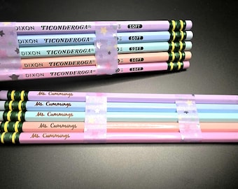 Puede incluir: Un conjunto de diez lápices de colores pastel dispuestos en dos filas, sujetos por cinta adhesiva translúcida con estampado de estrellas. Los lápices presentan el texto DIXON TICONDEROGA, SOFT y el nombre personalizado Ms. Cummings en cursiva.