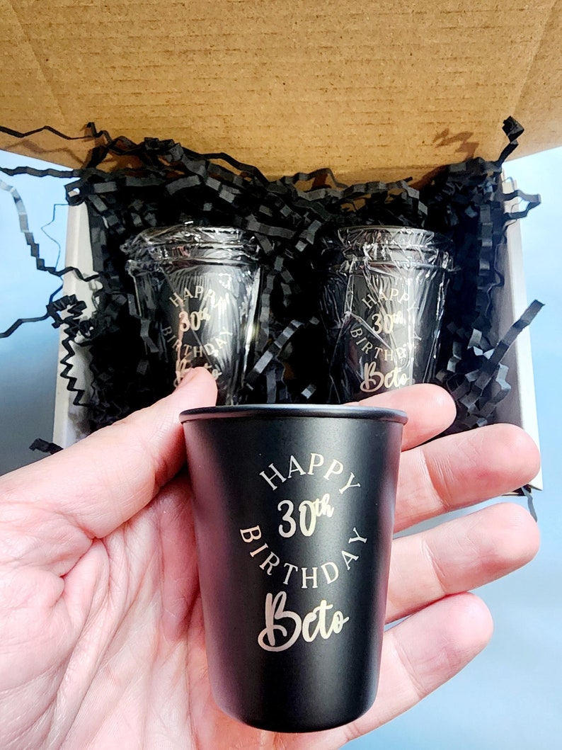 May include: Two black shot glasses with gold lettering that reads "Happy 30th Birthday Beto" in a circle. The shot glasses are in a box with black shredded paper.