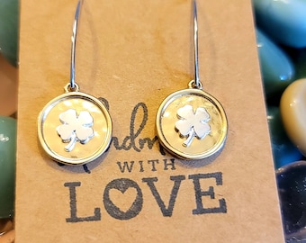 Puede incluir: Un par de pendientes colgantes de plata con un dije redondo dorado con un diseño de trébol de cuatro hojas. Los pendientes cuelgan de una tarjeta marrón con el texto "Made with Love".
