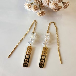 May include: Gold-toned threader earrings featuring a rectangular charm with a moon phase design. The earrings have a chain and are adorned with clear, faceted beads. The earrings are displayed on a white surface with floral accents.