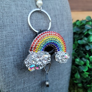 Puede incluir: Un portatarjetas con un dise&ntilde;o de arco&iacute;ris con un anillo plateado y una perla. El arco&iacute;ris est&aacute; hecho de pedrer&iacute;a de colores y las nubes est&aacute;n adornadas con pedrer&iacute;a transparente. El portatarjetas est&aacute; sujeto a un cord&oacute;n retr&aacute;ctil.