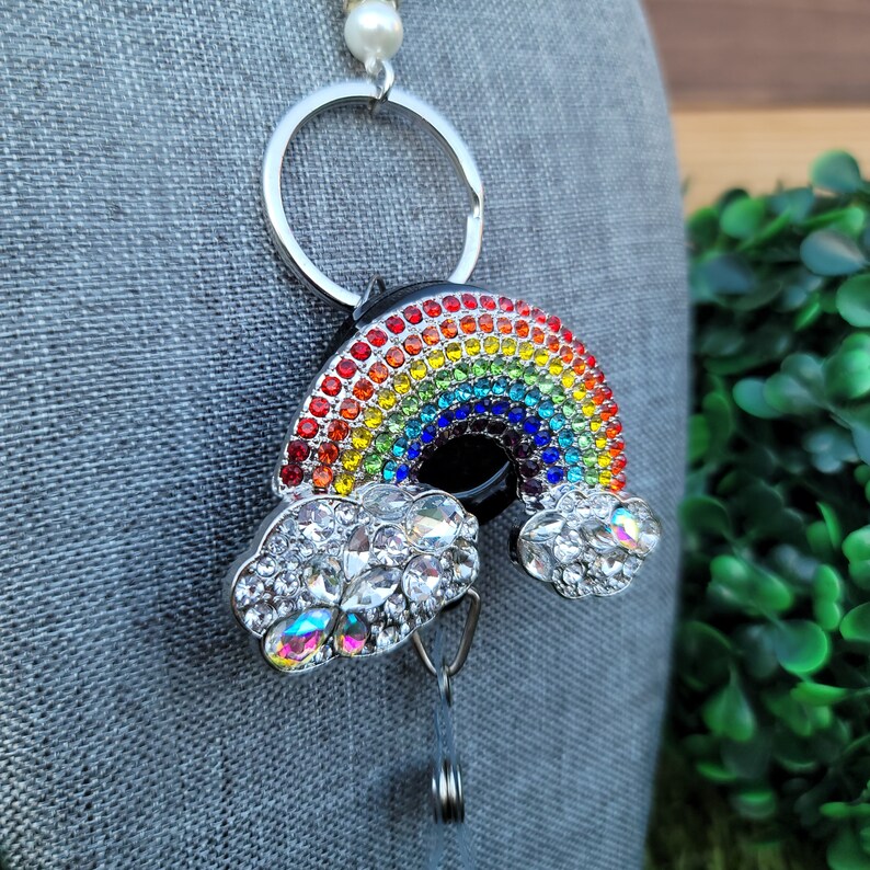 Puede incluir: Un portatarjetas con un dise&ntilde;o de arco&iacute;ris con un anillo plateado y una perla. El arco&iacute;ris est&aacute; hecho de pedrer&iacute;a de colores y las nubes est&aacute;n adornadas con pedrer&iacute;a transparente. El portatarjetas est&aacute; sujeto a un cord&oacute;n retr&aacute;ctil.