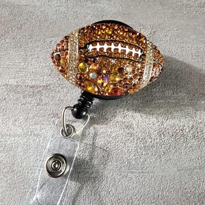 May include: A bedazzled football-shaped retractable badge holder. The football is covered in brown, gold, and clear rhinestones, with white stitching detail. The badge reel has a black base and a clear plastic strap with a metal snap.