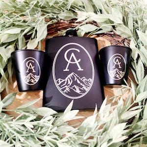May include: Black metal flask with a silver monogram "CA" and a mountain range design. Two matching black shot glasses with the same design.