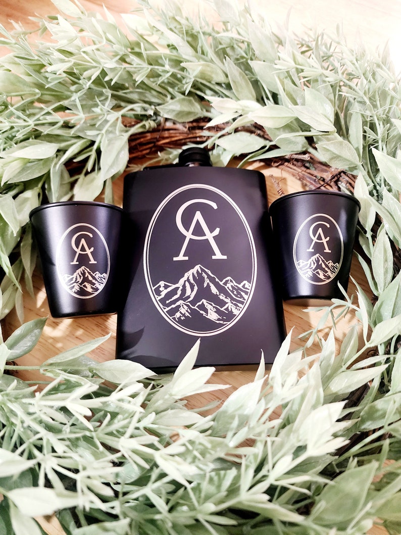 May include: Black metal flask with a silver monogram "CA" and a mountain range design. Two matching black shot glasses with the same design.