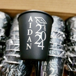 May include: Black plastic shot glass with the name "AIDAN" and the year "2024" printed in silver, along with a graduation cap graphic.