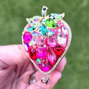 May include: A bedazzled strawberry-shaped badge reel. The pink base is covered in various sizes and shapes of colorful rhinestones. The stem and leaves are gold-toned with green and clear rhinestones. A silver clip and retractable cord are visible.