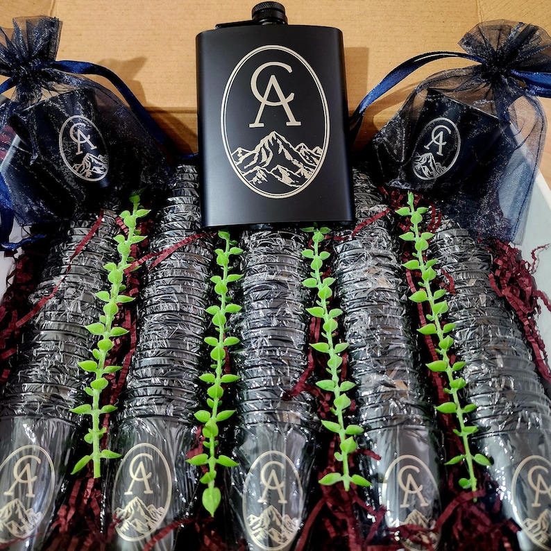 May include: A black flask with a gold engraved mountain and the initials "CA". The flask is surrounded by a variety of small black cups with the same gold engraving. The cups are arranged in rows and are surrounded by green and red decorative filler.