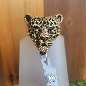 May include: A gold and black leopard head badge reel with rhinestones. The reel is attached to a clear plastic badge holder with a silver clip.