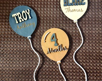 May include: Three wooden balloon-shaped decorations. One is blue with "TROY" and "Anthony" in white and gold lettering. Another is cream-colored with "BLAKE" and "Thomas" in blue and gold. The third is tan with "4 Months" in blue and gold.