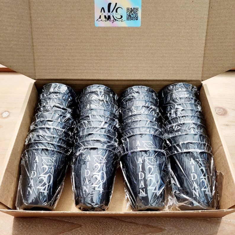 May include: A cardboard box filled with black plastic cups. Each cup has the text "Aidan 2024" printed on it.