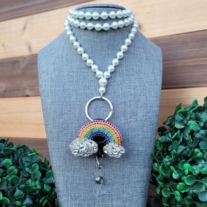 Puede incluir: Un collar de cord&oacute;n de cuentas blancas con un portatarjetas en forma de arco&iacute;ris. El arco&iacute;ris est&aacute; incrustado con pedrer&iacute;a de colores y tiene dos acentos de nubes con pedrer&iacute;a. El portatarjetas tiene un carrete retr&aacute;ctil y un anillo plateado.