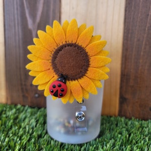 May include: A felt sunflower with a brown center and a red ladybug on the front. The sunflower is attached to a clear plastic badge holder.