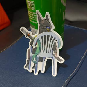 Puede incluir: Una pegatina troquelada que presenta un personaje de estilo anime con una cabeza negra y una espada, sentado en una silla de plástico blanca. El personaje tiene el pelo verde y lleva un atuendo amarillo y azul. La pegatina está delante de una lata verde.
