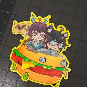 May include: A die-cut sticker featuring two anime-style characters with dark hair riding inside a large orange burger-shaped vehicle. One character has small horns and a flag with the text ENDFIELD, while the other wears red-rimmed goggles.