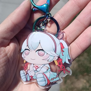 May include: A clear acrylic keychain featuring a cartoon character with white hair, pink eyes, and a light blue outfit. The keychain has a colorful iridescent clasp and ring. The character is sitting with its legs crossed and has red accents.