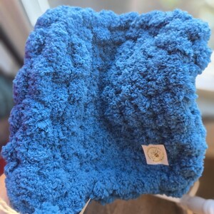 May include: A square, plush, blue blanket with a textured surface. The blanket has a small tag with a bee and the words "Bumble Bee Knits". The blanket is made of a soft, fluffy material.