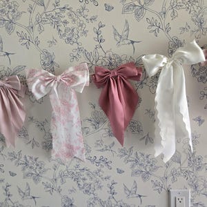 May include: A horizontal garland featuring five decorative fabric bows in varying shades of pink, white, and cream. The bows include solid satin textures, a white and pink floral toile print, and a light pink and white gingham check pattern.