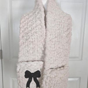 May include: A light pink, chunky knit scarf with two pockets and a black bow accent. The scarf is displayed on a white mannequin head against a white door background. The scarf is made of a soft, textured material.