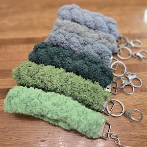 May include: A collection of fluffy keychains in various shades of green and blue, arranged on a wooden surface. Each keychain features a soft, textured fabric loop attached to a silver-colored metal clasp and keyring. The keychains are in a row, showcasing the different colors.