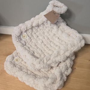 May include: Two off-white, plush pot holders with a textured, looped design. Each pot holder has a small, round button and a brown tag attached. The pot holders are folded and rest on a wooden surface.