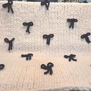 May include: A cream-colored, chunky knit blanket with several dark navy blue bow accents. The bows are evenly spaced across the surface of the blanket. The blanket is displayed on a surface.