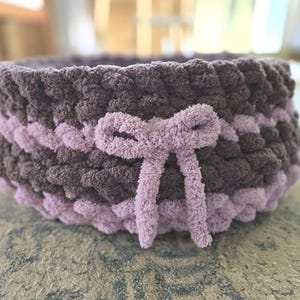 May include: A handmade, round basket in shades of brown and lavender. The basket is made of a thick, textured yarn, with a bow detail in the center. The basket is suitable for storage or decoration.