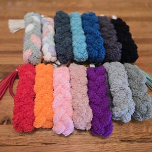 May include: A collection of braided keychains in various colors, including red, orange, pink, purple, gray, and black. Each keychain features a fluffy, textured braid and a coordinating tassel. The keychains are arranged on a wooden surface.