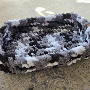 May include: A rectangular pet bed in a camouflage pattern of black, white, and gray. The bed is made of a soft, plush material, with a thick, raised edge. The bed is designed for small pets.