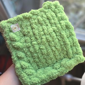 May include: A square, lime green, plush, knitted item. The item has a textured surface with vertical ridges. A small, rectangular tag with a bee logo and text is attached to the item.
