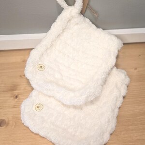 May include: Two off-white, fluffy pot holders with a loop for hanging. Each pot holder has a small, round label with a bee design. The pot holders are made of a soft, textured material and are sitting on a wooden surface.