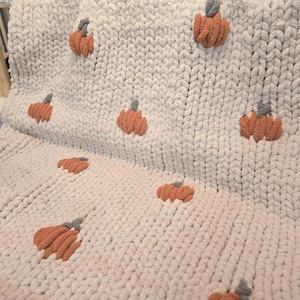 May include: A cream-colored, chunky knit blanket adorned with multiple orange and gray pumpkin appliqués. The blanket is folded over, showcasing the texture and design, perfect for fall decor.