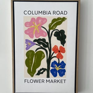 May include: A framed art print with a floral design and the text "COLUMBIA ROAD FLOWER MARKET." The artwork displays stylized flowers in pink, red, blue, and purple, with green leaves against a cream background. The frame is a light brown colour.