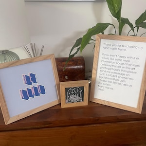 May include: Three framed artworks with wooden frames. One frame displays the words "BE KIND" in blue and pink. Another frame contains a small black and white illustration. The third frame has text that reads "Thank you for purchasing my hand made frame."