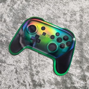 May include: A green, black, and iridescent sticker of a video game controller. The controller has buttons labeled A, B, X, and Y.