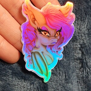 May include: Holographic sticker featuring a stylized fantasy creature with orange and pink hair, and a blue and purple face. The sticker has a pearlescent, iridescent finish, and a detailed illustration style.