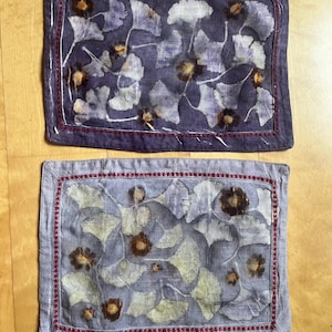 May include: Two rectangular fabric placemats featuring botanical prints of ginkgo leaves and circular flower heads. The top placemat is deep purple, while the bottom one is a lighter lavender, both bordered by a decorative red stitched frame.