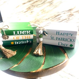 May include: Two stacked wooden books with a green, white, and gold color scheme. The top book reads "LUCK OF THE IRISH" and the bottom book reads "HAPPY ST. PATRICKS DAY". Both books have a green shamrock and a brown tassel.