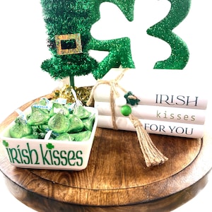 May include: A wooden round tray with a green shamrock, a green leprechaun hat, and a stack of books that say "IRISH kisses FOR YOU". A white bowl filled with green chocolate kisses says "Irish kisses" on the front.