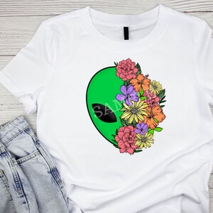May include: White t-shirt with a green alien head graphic and a floral design. The flowers are pink, yellow, orange, and purple.