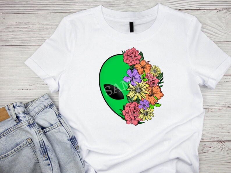 May include: White t-shirt with a green alien head graphic and a floral design. The flowers are pink, yellow, orange, and purple.