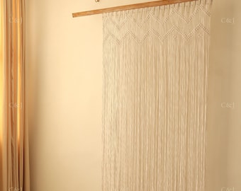 May include: A cream-colored macrame wall hanging suspended from a horizontal wooden rod. The piece features a series of intricate knotted chevron patterns at the top, transitioning into long, straight, vertical fringe that hangs downward.