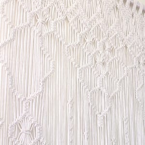 Könnte beinhalten: Weißer Makramee-Wandbehang mit geometrischem Design. Das handgefertigte Textil weist komplizierte Knotenmuster auf, die ein strukturiertes und dekoratives Stück schaffen. Die Wanddekoration ist an einem Holzstab aufgehängt und verleiht jedem Raum einen Hauch von Bohème-Stil.