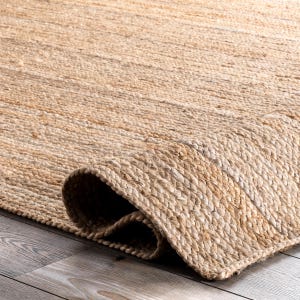 May include: A natural fiber rug with a rolled-up edge. The rug is made of woven jute fibers in a light tan color. The texture of the rug is visible, with a close-up view of the woven pattern. The rug is on a wooden floor.