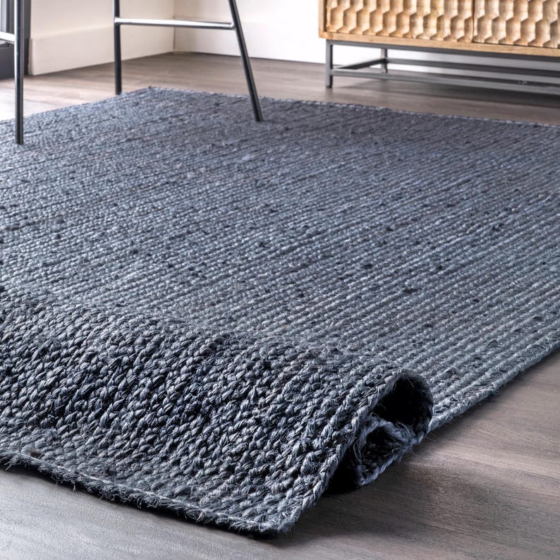May include: A rectangular, dark gray jute rug with a rolled-up edge. The rug has a woven texture and is placed on a wooden floor. The rug's color is a deep, neutral gray, suitable for various interior styles.