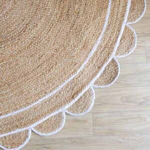 May include: A round jute rug with a scalloped edge and a white border. The rug has a natural, woven texture and a light brown color. The white border adds a contrasting element to the design. The rug is placed on a light wood floor.