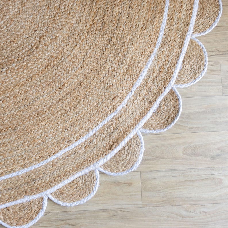 May include: A round jute rug with a scalloped edge and a white border. The rug has a natural, woven texture and a light brown color. The white border adds a contrasting element to the design. The rug is placed on a light wood floor.