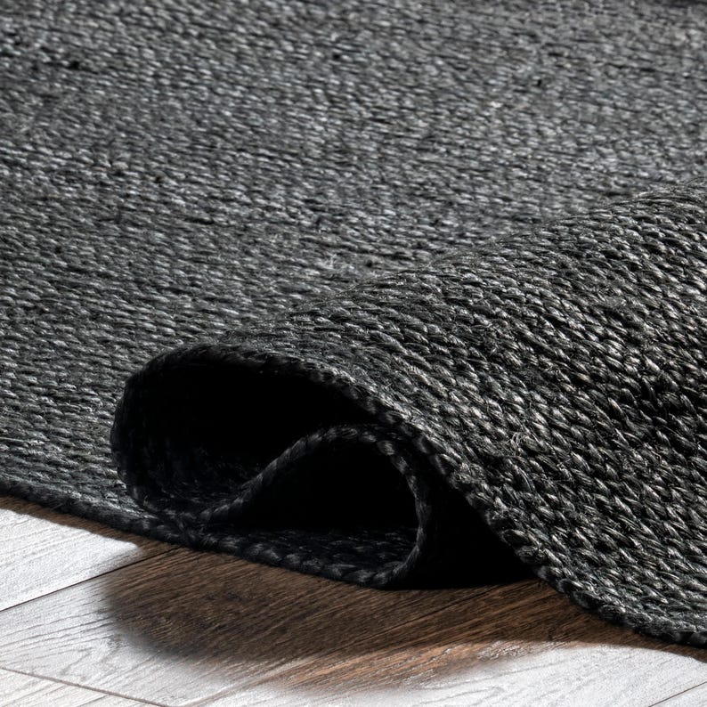 May include: A rolled-up, dark gray woven rug. The rug's texture is visible, with a close-up view highlighting the woven fibers. The rug is on a wooden surface, with the rolled edge creating a shadow.